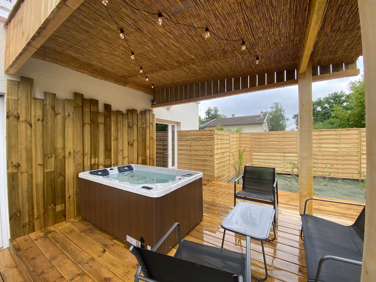 A private outdoor area features a hot tub sheltered by a pergola, surrounded by wooden fencing for added privacy. String lights provide soft illumination, and outdoor furniture including chairs and a table is arranged on a wooden deck, inviting relaxation.