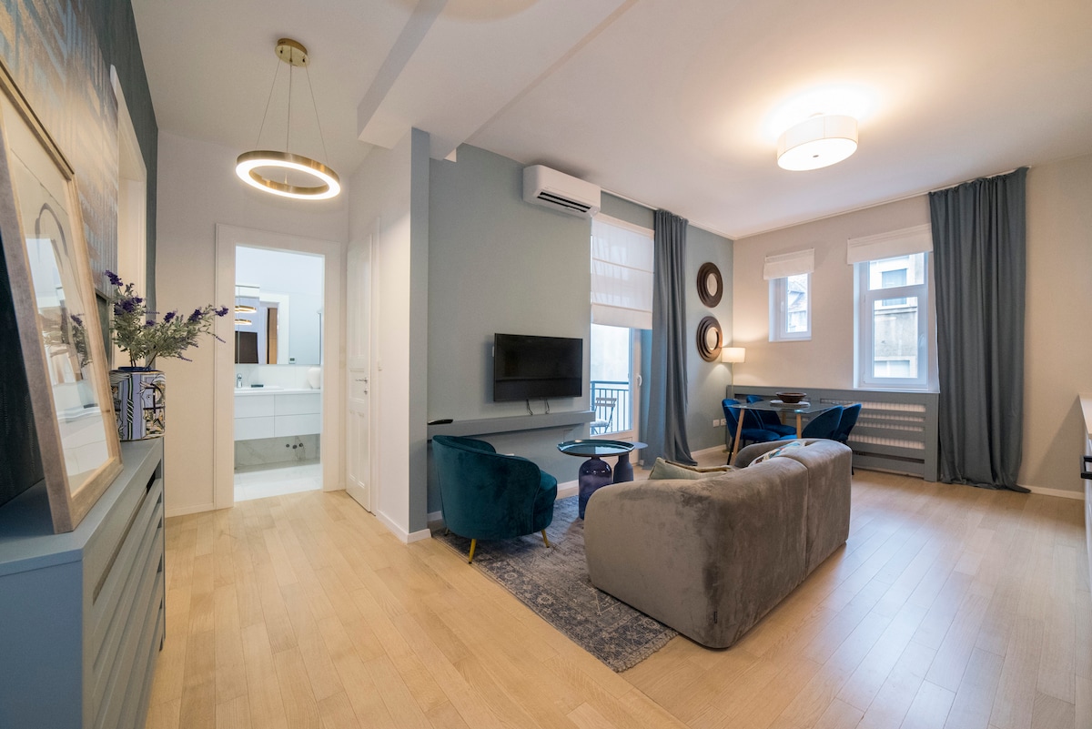 A spacious living area is presented with a comfortable sofa, accent chairs, and a decorative table. Natural light enters through large windows covered by sheer curtains. A television is mounted on the wall, and a doorway leads to a bathroom, visible in the background.