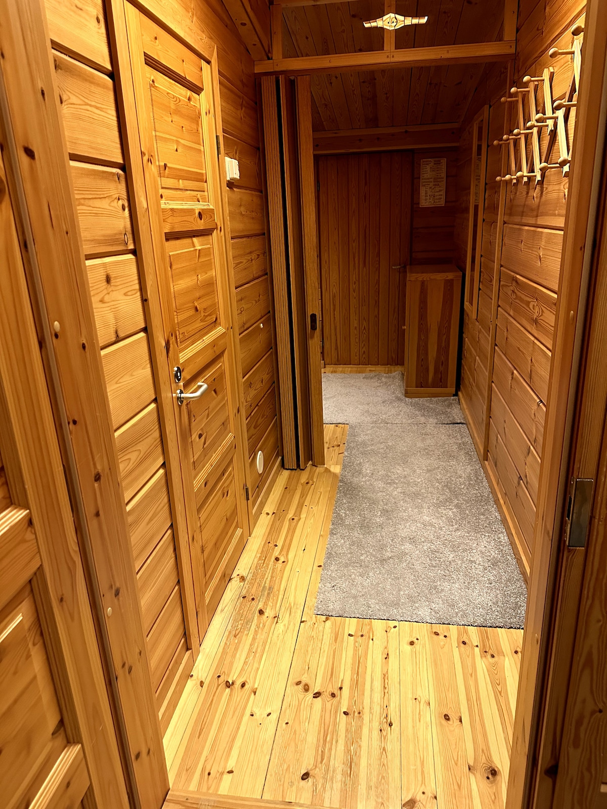 A hallway is presented with wood paneling and warm-toned wooden floors. The space features two closed doors on the left, and a spacious area is visible at the end. A grey carpet runner provides contrast against the wood, enhancing the cozy ambiance.