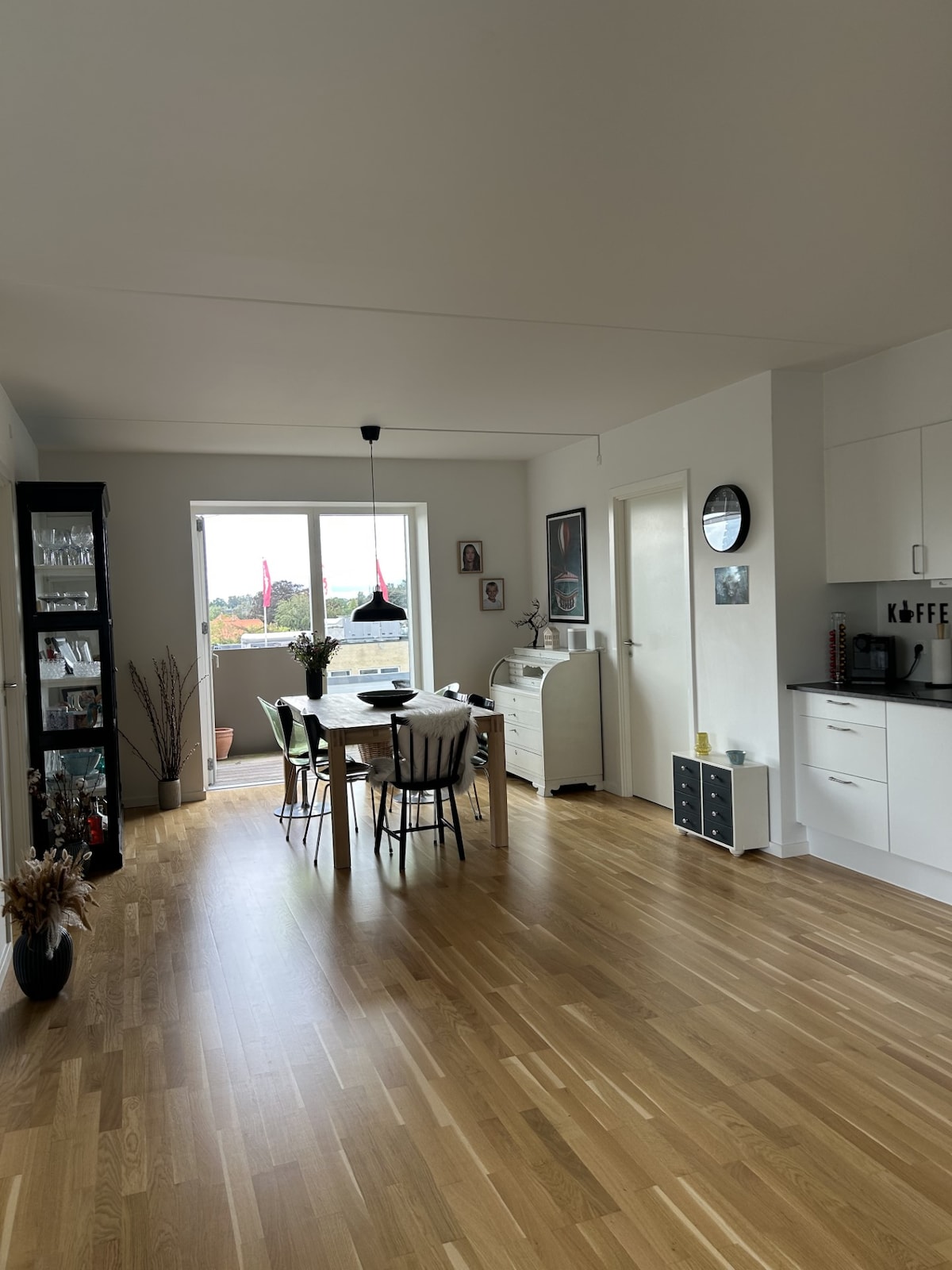 A light-filled dining area features a wooden table surrounded by six chairs. A modern pendant light hangs above the table. Cabinetry and appliances are visible in the kitchen area, which transitions into the dining space. Large windows invite natural light and provide views of the outside.