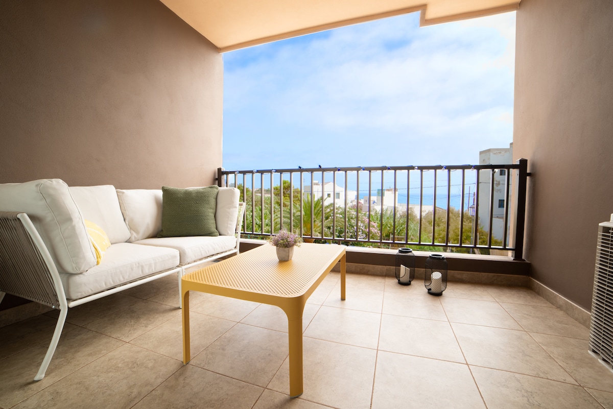 A balcony area is presented with a light-colored sofa accompanied by a green cushion. A yellow coffee table is positioned in front of the sofa. In the background, the ocean can be seen through the railing, with greenery and buildings softly visible.