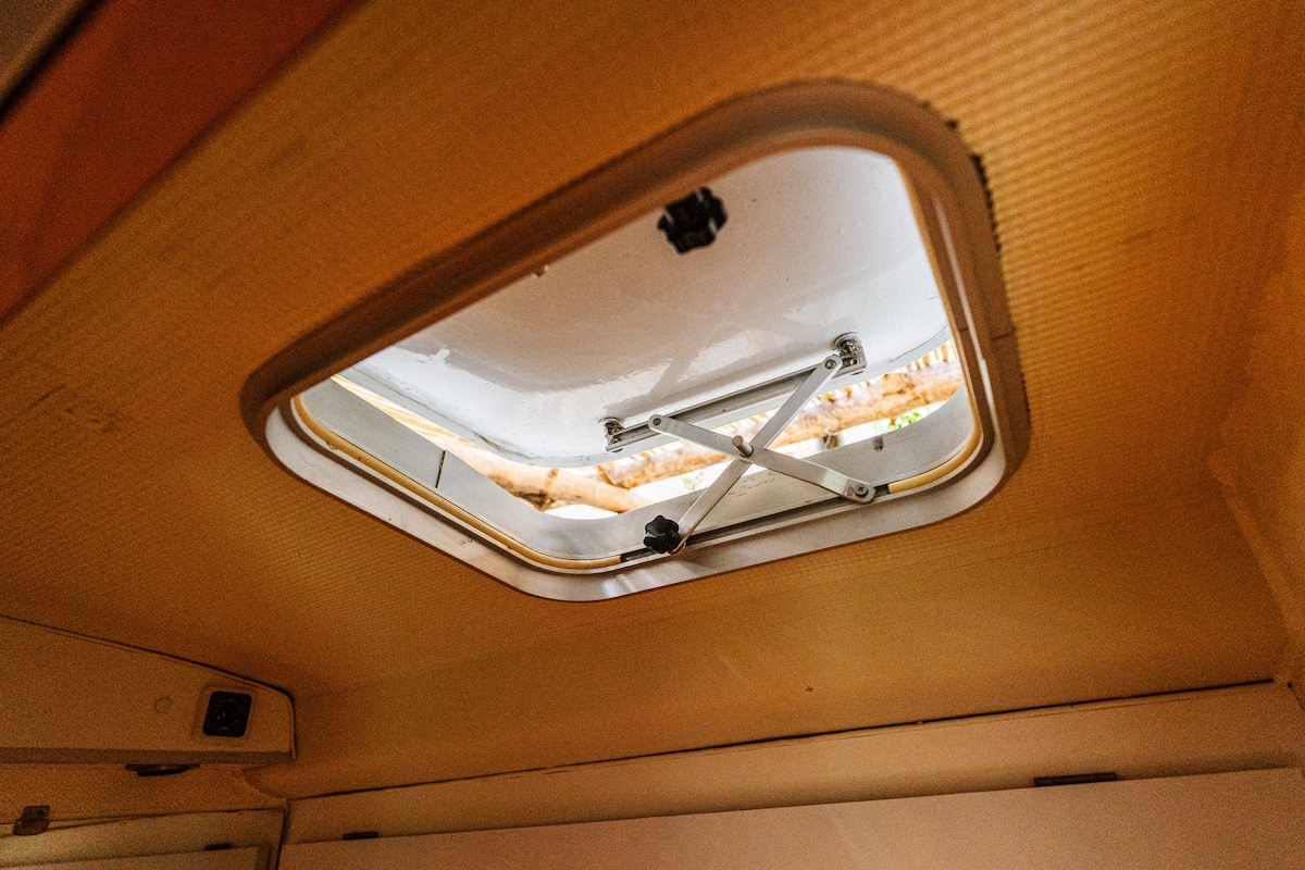 A ceiling hatch is visible, featuring a white frame and a mechanical opening mechanism. Natural light filters through the hatch, hinting at the greenery outside, while the surrounding interior offers a sense of spaciousness and connection to the outdoor environment.