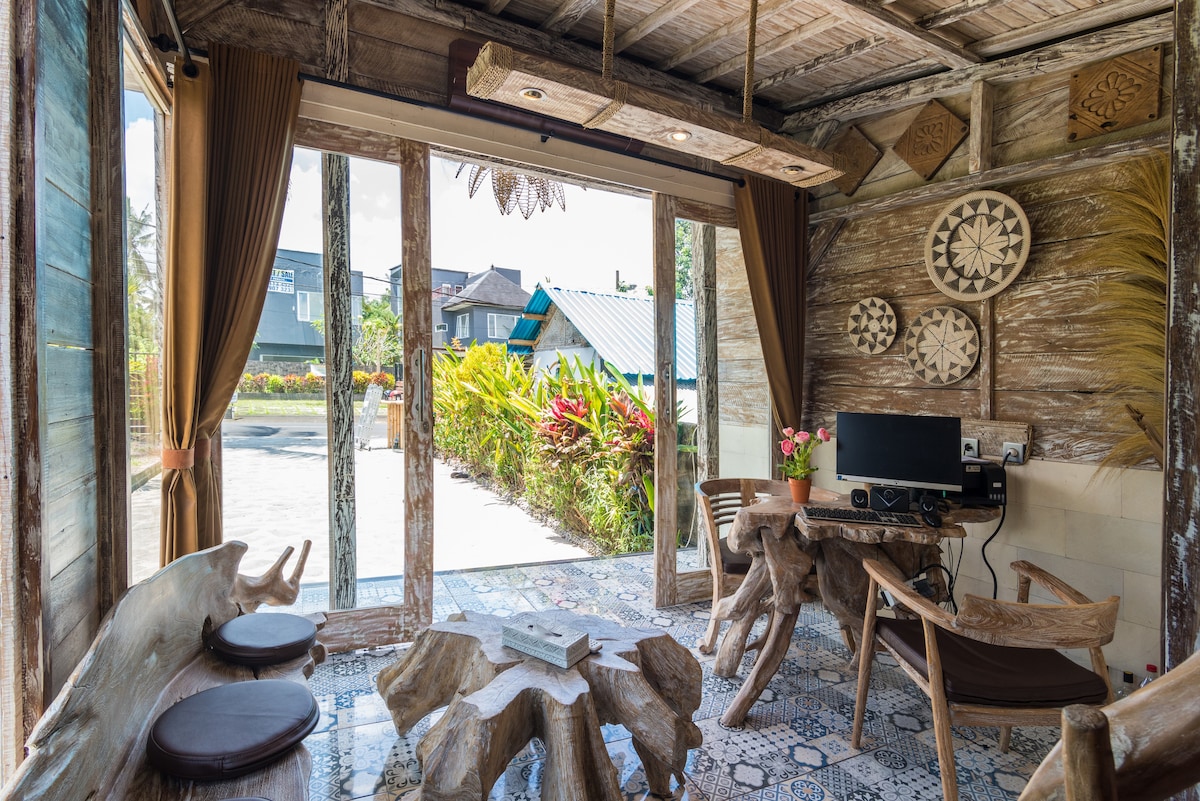 A cozy living area features rustic wooden furniture, including a unique table and seating crafted from natural wood. Large glass doors provide a view of the vibrant garden and surroundings. Decorative wall art consisting of intricate carvings adds character to the space.