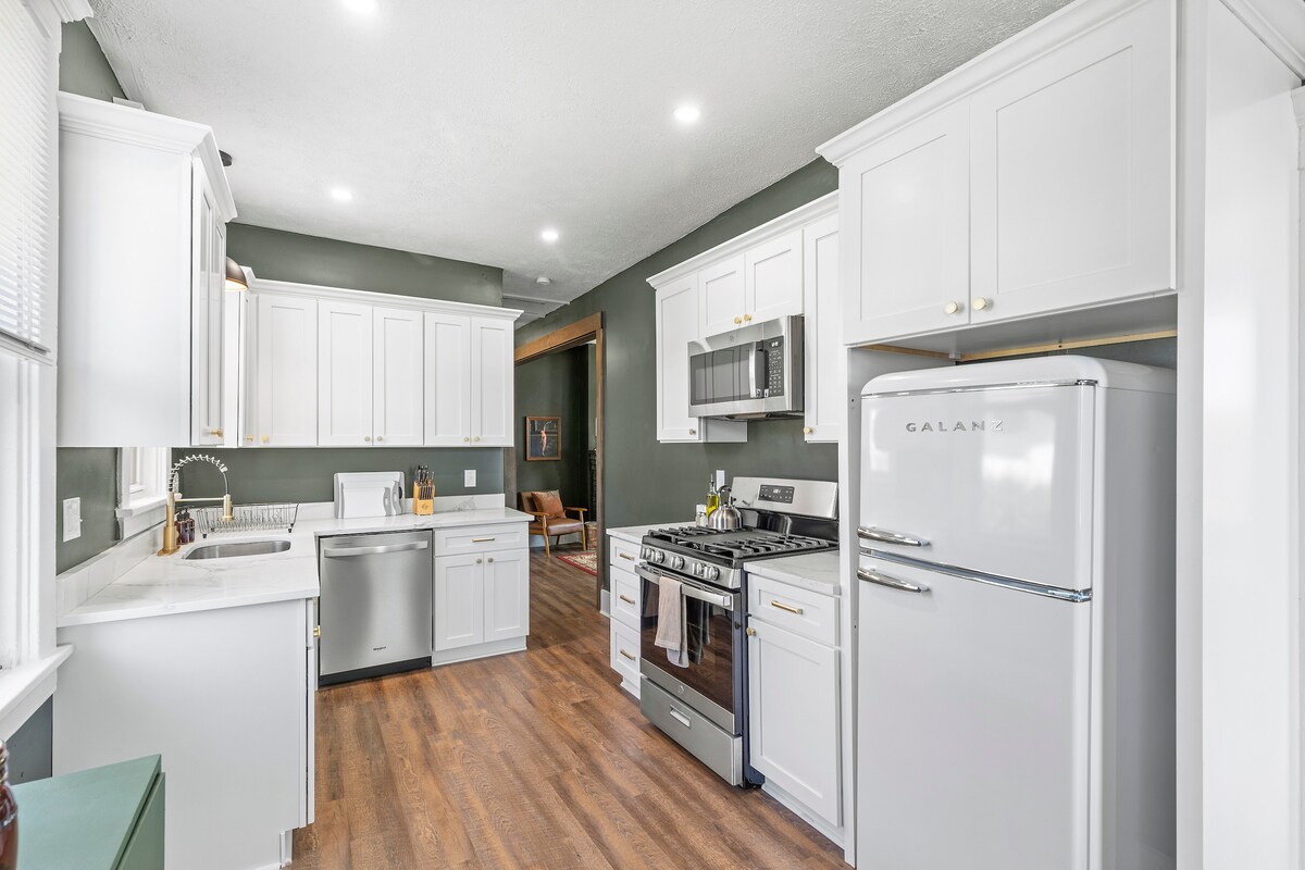 The modern kitchen features white cabinetry with gold accents and stainless steel appliances, including a refrigerator, stove, and dishwasher. A large window allows natural light into the space, enhancing its bright and inviting design. The countertops are smooth and ample for meal preparation.
