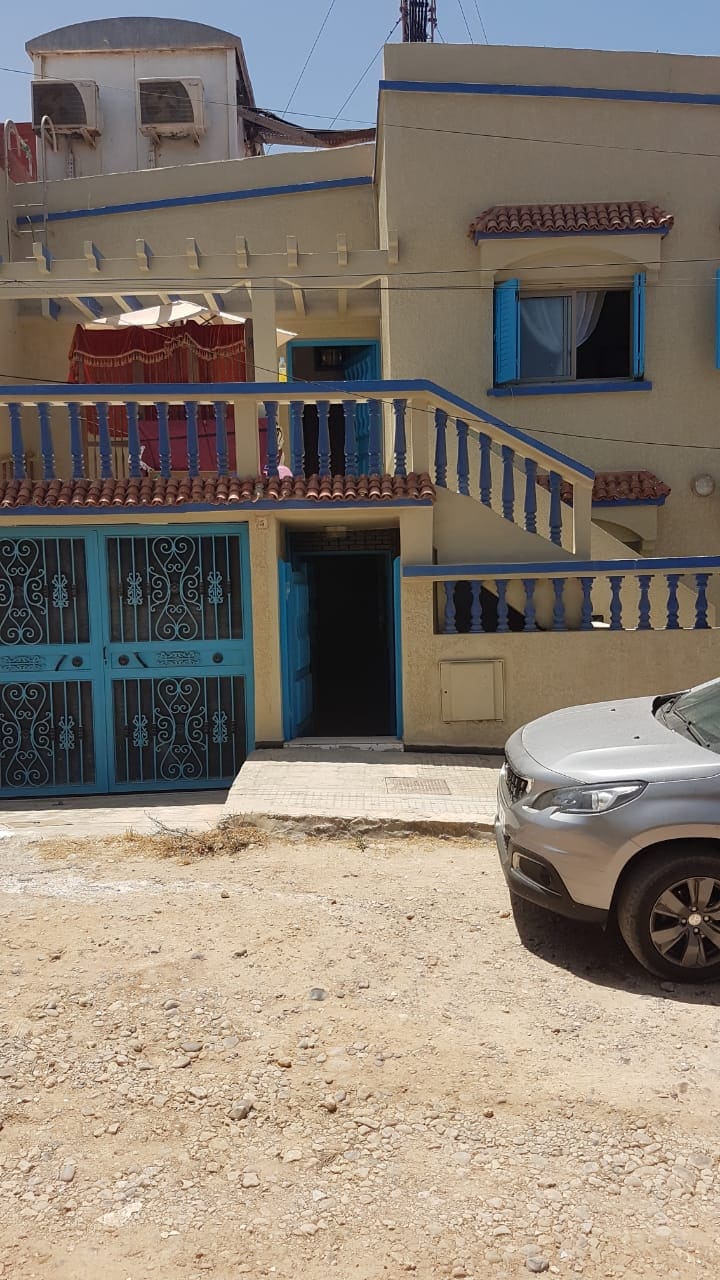 The exterior of a two-story building is visible, featuring a light-colored facade and blue railings. The entrance is marked by a solid door, with open windows adorned by light curtains. A parked vehicle is positioned in the foreground on a gravel surface.
