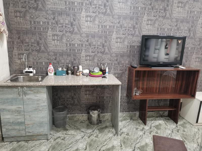 A compact kitchenette is displayed with a granite countertop and a small sink. Various dishes and utensils are organized along with cleaning supplies. A television is mounted on a wooden stand nearby, while a trash bin is positioned under the counter.