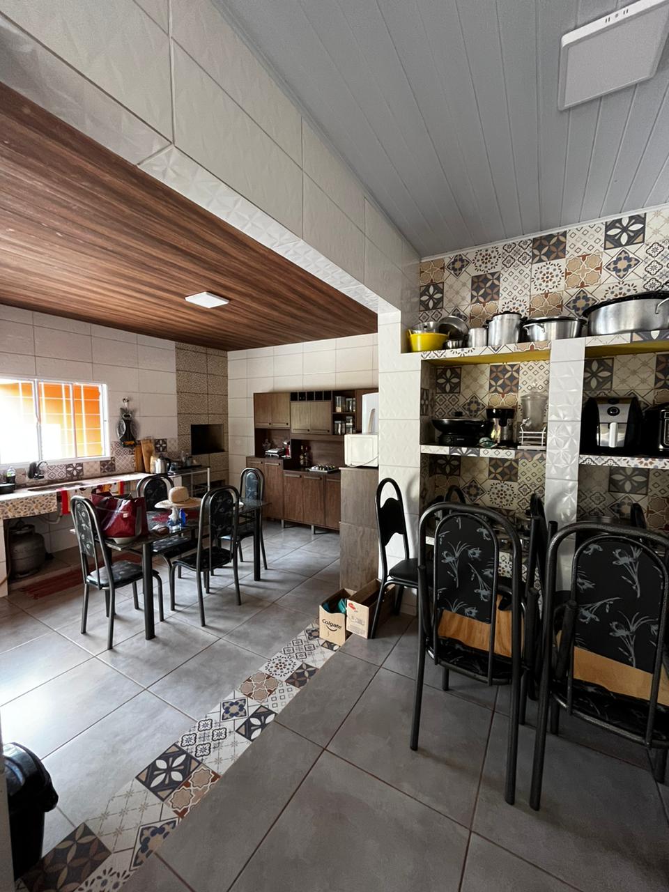 The kitchen area features a blend of tiled walls and wooden accents, with an arranged dining table and chairs. Appliances, including coffee machines and a microwave, are organized on shelves. Natural light enters through a window, illuminating the space.