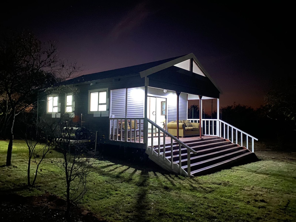 The cabin is illuminated at night, highlighting its welcoming front porch and steps. Soft lighting spills from the large windows, offering a glimpse of the comfortable seating inside. The surrounding grass is neatly maintained, creating an inviting atmosphere amidst the natural landscape.