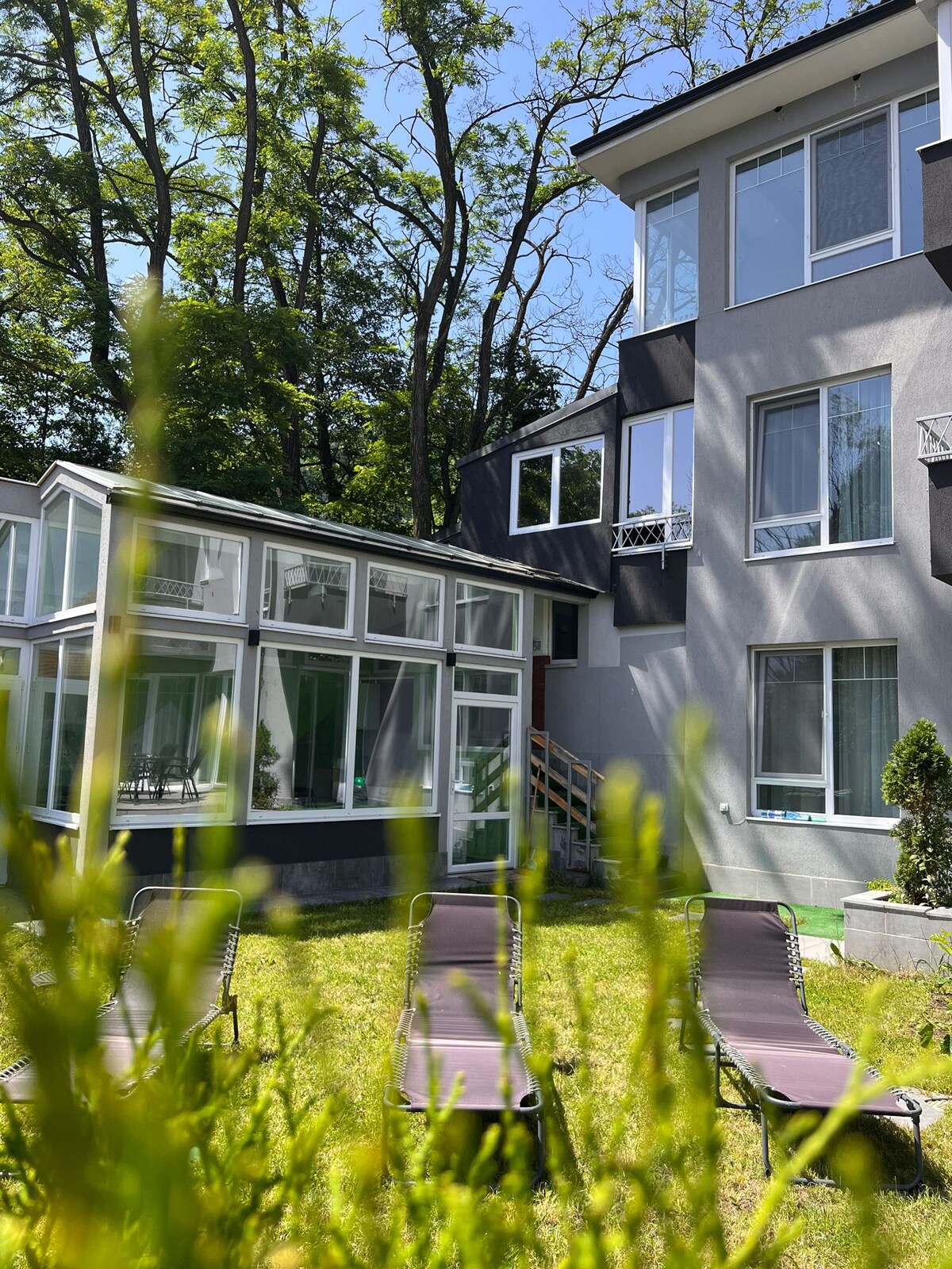 A portion of the villa is captured, showcasing its modern architecture against a backdrop of trees. Several lounge chairs are positioned on the lush green lawn, while a glass-enclosed area is visible, offering sunlight and a connection to the outdoors.