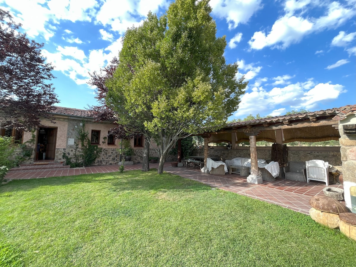 A spacious outdoor area features a covered porch with multiple seating arrangements, surrounded by lush greenery and vibrant trees. The rustic stone house is visible in the background, under a blue sky with scattered clouds.