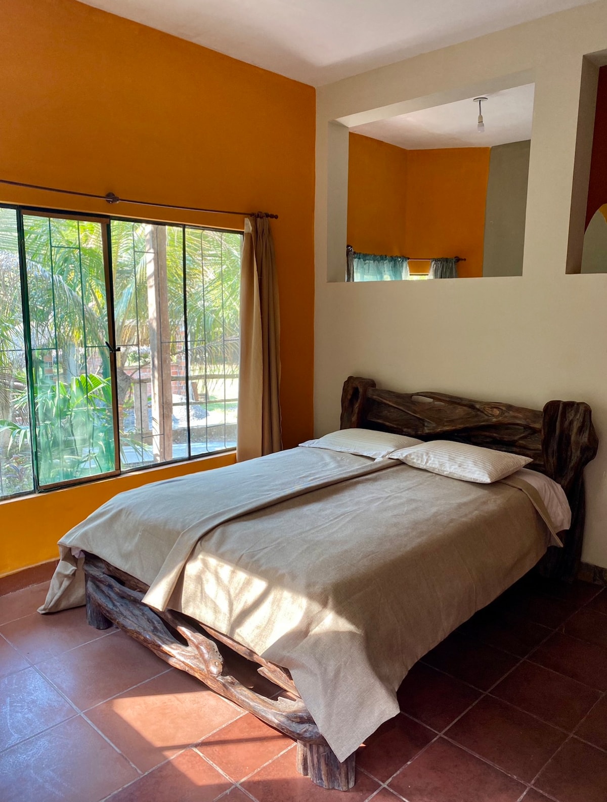 A cozy bedroom features a wooden bed with a neutral-toned bedding set. A large window allows natural light to fill the space, framing views of lush greenery outside. The walls are painted in warm shades of orange and beige, contributing to a welcoming ambiance.