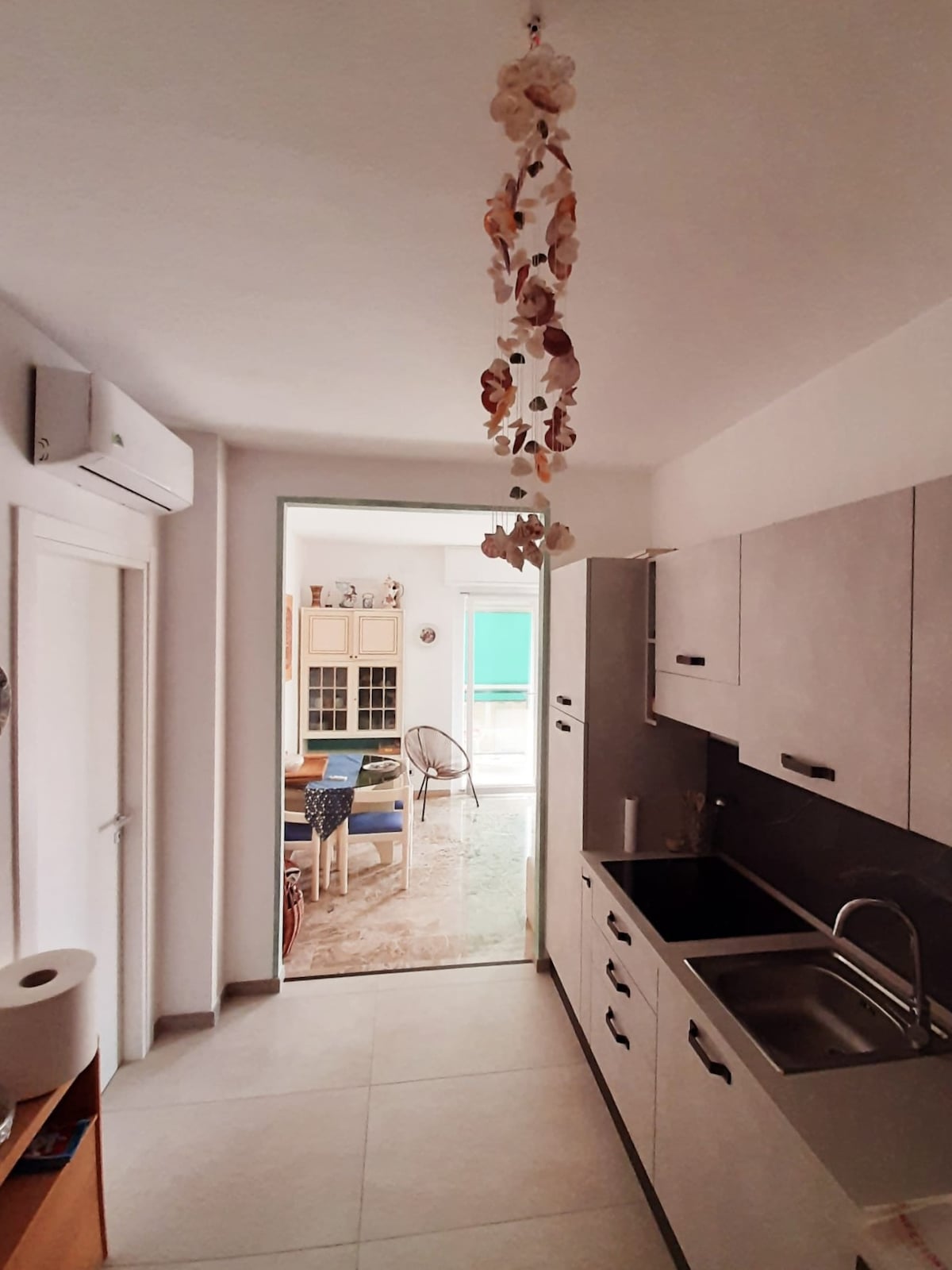 A modern kitchen area is shown with sleek cabinetry and a stainless steel sink. Natural light flows in from an open doorway leading to a bright living space. Decorative elements hang from the ceiling, creating a welcoming ambiance.