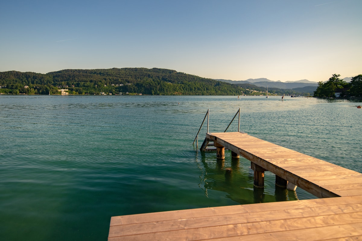 A wooden dock extends into calm waters, providing access to the lake. The surrounding landscape features gently rolling hills and green trees, while distant mountains rise in the background. The clear sky reflects a soft golden light on the water's surface.