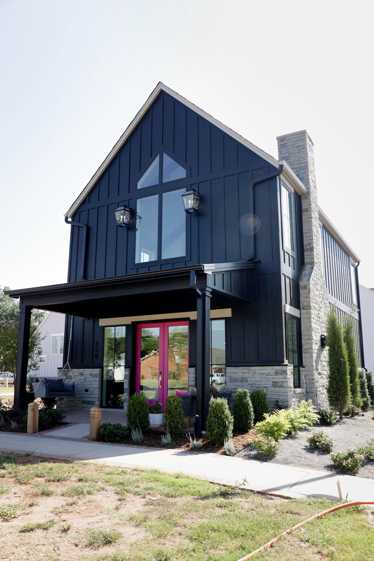 A modern two-story home features a bold black exterior with contrasting stone accents. Large windows allow natural light to fill the interior, while a pink door adds a vibrant touch to the entrance. Lush greenery surrounds the path leading to the home.