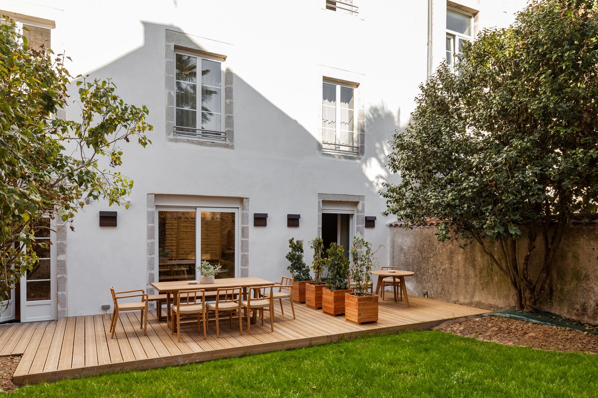 A spacious wooden deck extends into a lush green garden area, featuring a dining table surrounded by chairs. Potted plants add a touch of greenery, while the exterior wall showcases large windows with a simple, modern design.