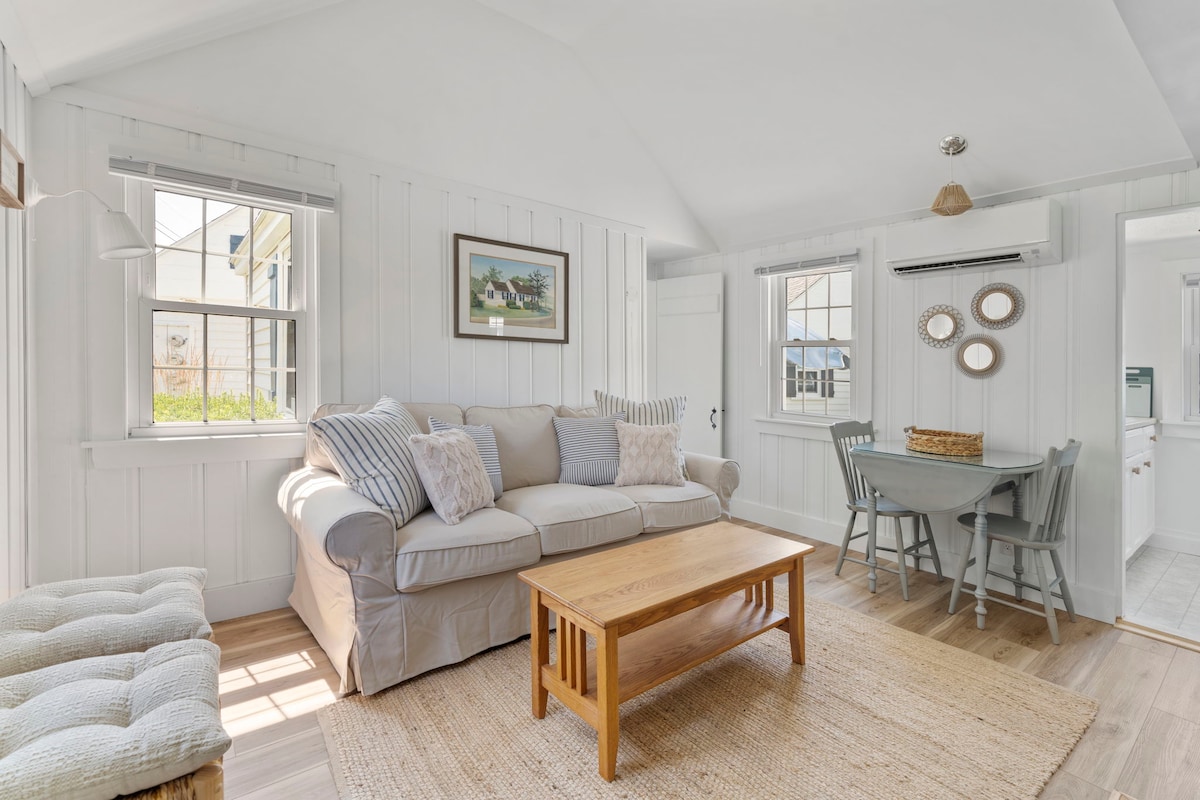 The living area features a light-colored sofa adorned with decorative pillows, paired with a wooden coffee table. A round dining table with chairs is visible nearby. Natural light filters through multiple windows, highlighting the white shiplap walls and calming atmosphere.