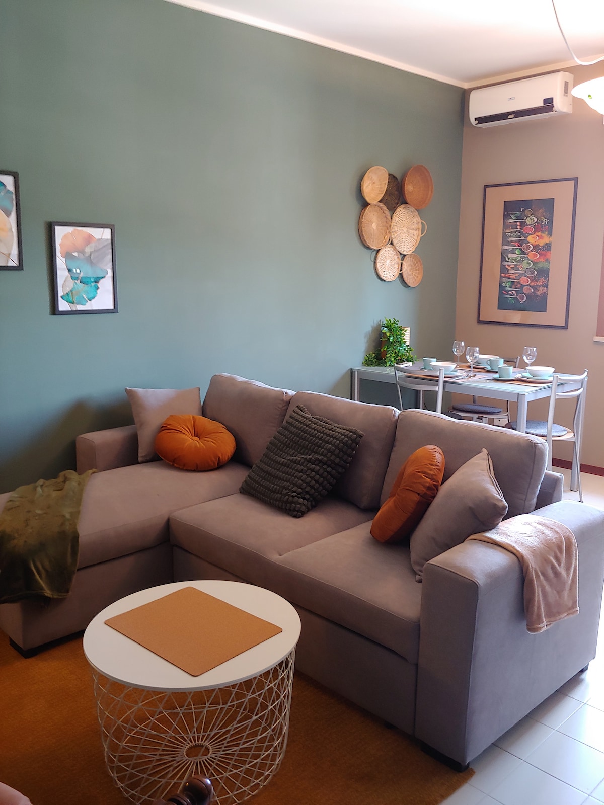 A cozy living area features a sectional sofa adorned with colorful cushions and a throw. A round coffee table sits in front, while a dining table is set for four in the background. Soft wall art and decorative plates add character to the green walls.