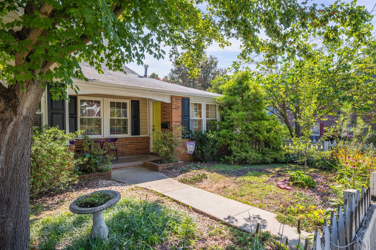 The inviting exterior of the residence features a welcoming front porch with seating, surrounded by a well-kept garden. Lush greenery and a birdbath enhance the tranquil atmosphere, with sunlight filtering through the trees, creating a peaceful outdoor escape.