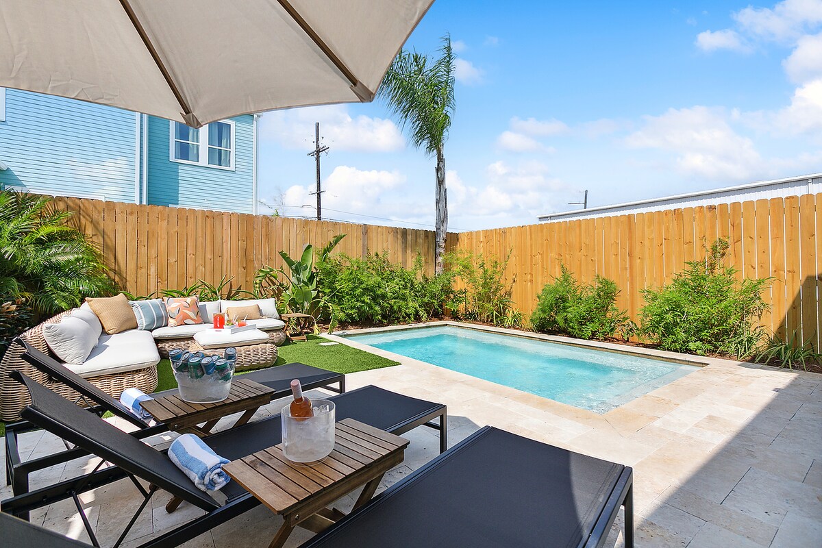 An outdoor space is highlighted by a private heated pool surrounded by tropical plants. Comfortable lounge chairs and a low table are arranged neatly on the patio, inviting relaxation. A large umbrella provides shade, enhancing the tranquil setting.