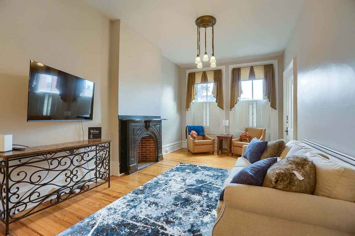 A spacious living area features a modern television mounted on the wall, complemented by two cozy armchairs. A decorative fireplace provides an attractive focal point, while large windows allow natural light to fill the room. A soft area rug adds texture to the hardwood floor.