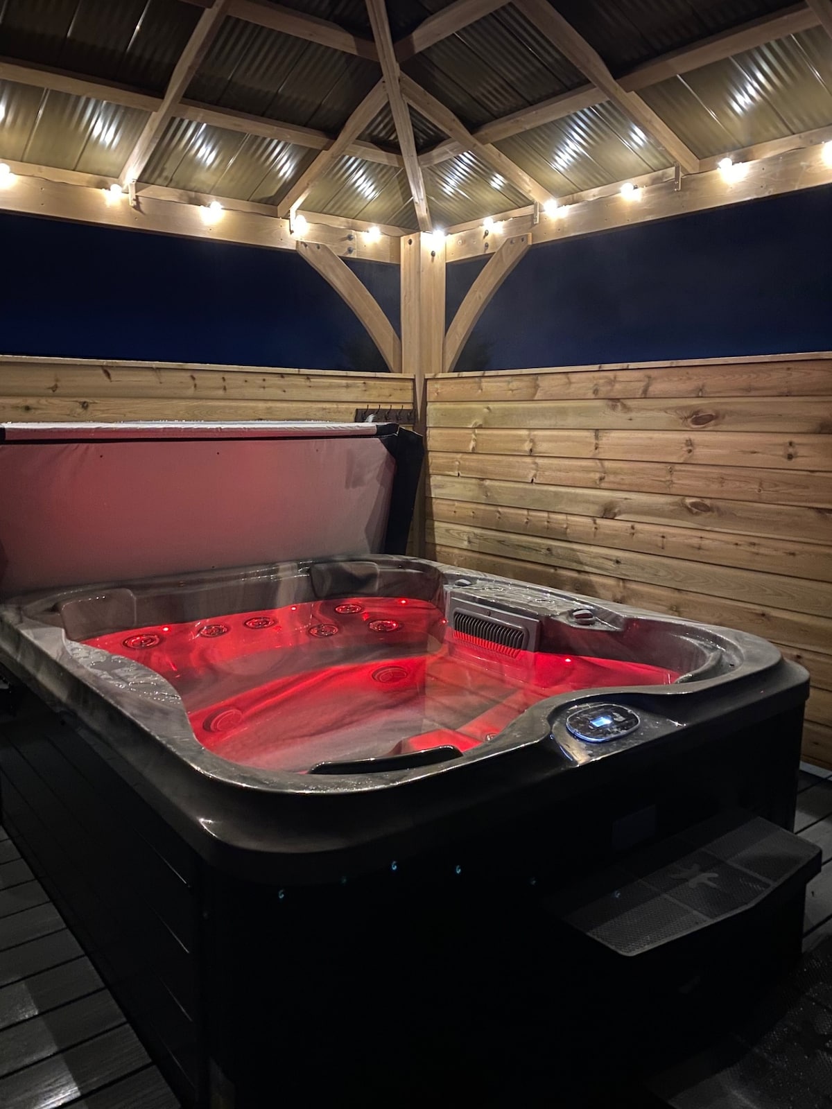 A hot tub is positioned under a wooden structure with overhead lighting, creating a relaxing ambiance. The tub is filled with water that reflects red illumination, highlighting its spacious design. Wooden walls provide privacy while maintaining an open feel.