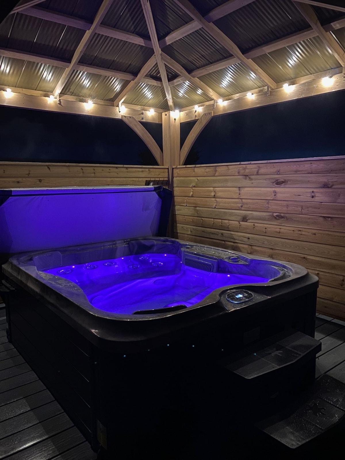 A hot tub is situated beneath a wooden gazebo, featuring warm lighting around its perimeter. The tub displays soft purple hues, creating a calming ambiance. Natural wood walls encase the area, providing privacy while complementing the overall design.