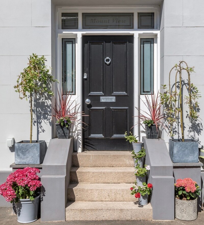 The entrance features a dark front door adorned with a circular knocker. Flanking the steps are decorative planters filled with vibrant flowers, including pink geraniums and green plants. The stone pathway leads up to the entrance, providing an inviting initial view of the property.