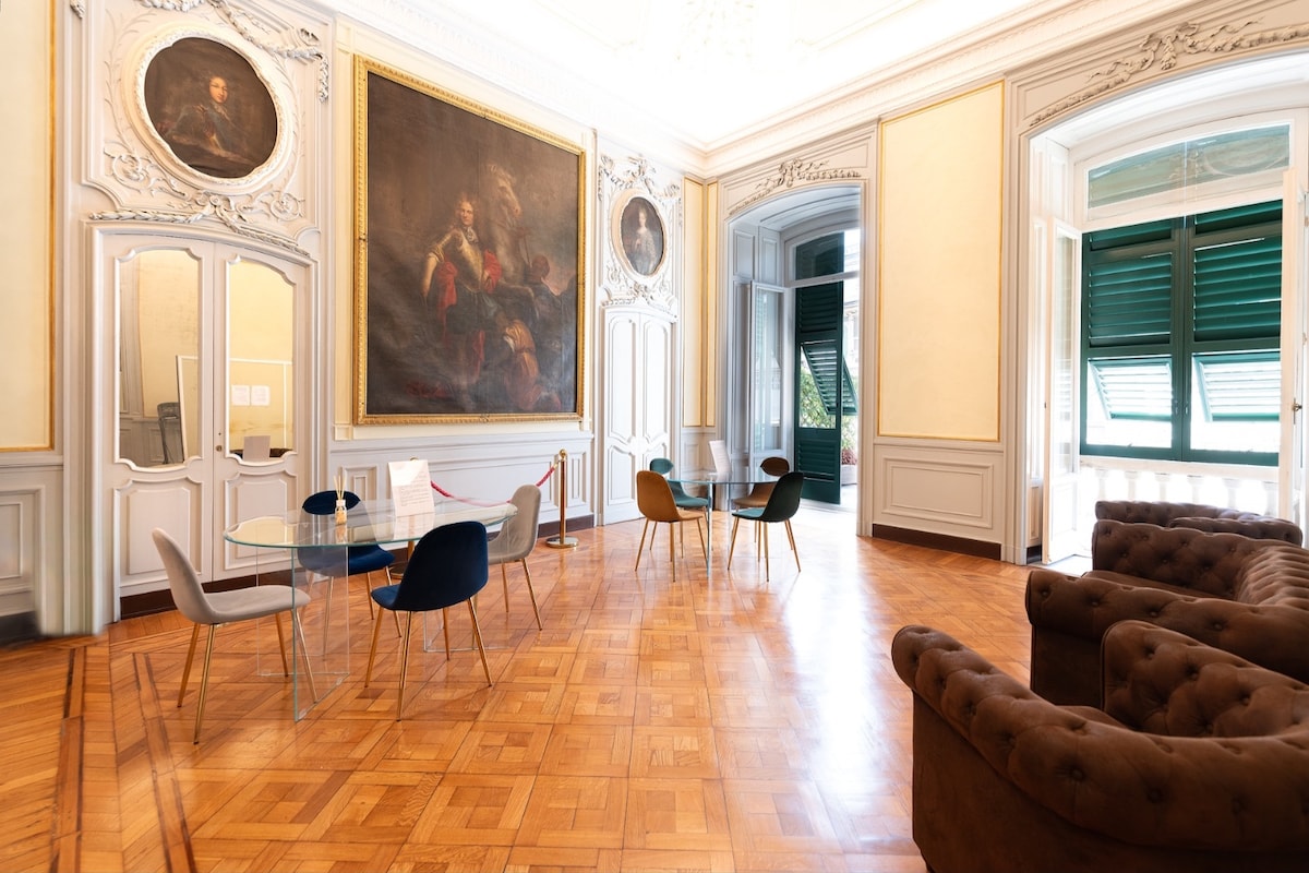 An elegant communal area features a wooden floor and high ceilings adorned with intricate moldings. An oversized painting is displayed prominently on the wall, surrounded by classical portraits. A glass dining table is accompanied by colorful chairs, while a plush brown sofa offers a cozy seating option.