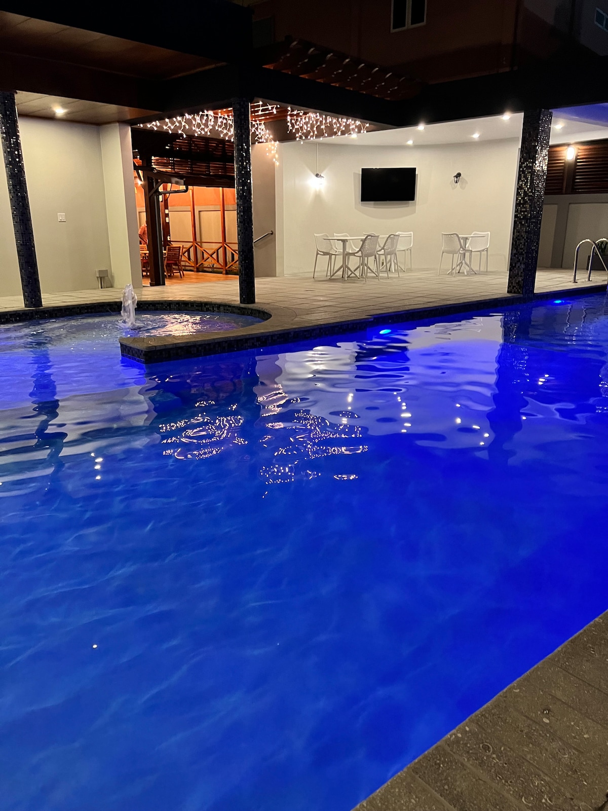 A well-lit swimming pool features vibrant blue water, reflecting the surrounding lights. An outdoor dining area with white furniture is visible in the background, along with a wall-mounted television. Water features add a calming element, enhancing the serene environment of the space.
