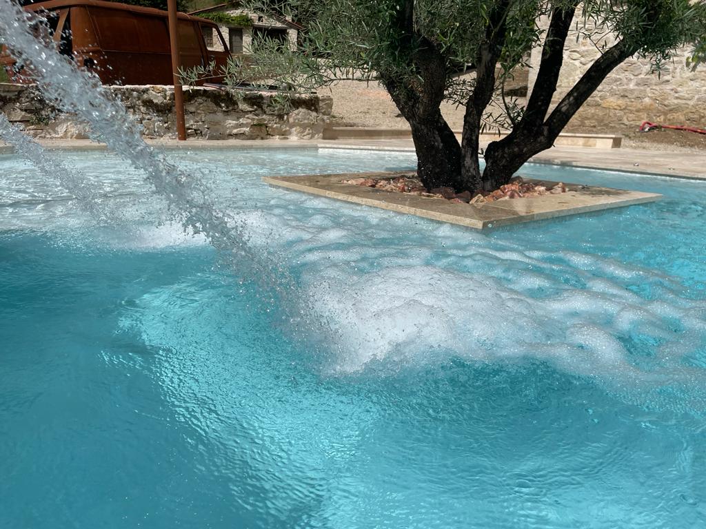 The image showcases a clear blue pool with bubbles gently emerging from a submerged fountain feature. In the center, a small island displays an olive tree surrounded by pebbles, adding a touch of nature to the tranquil scene.