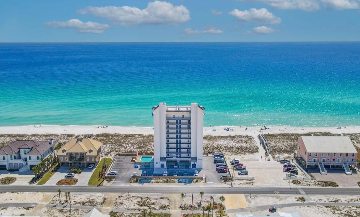 An aerial view showcases a modern building situated near the beach, with soft white sands and vibrant turquoise waters in proximity. The beach is visible, with a few people enjoying the sunshine. Nearby structures and ample parking spaces are also present in the foreground.