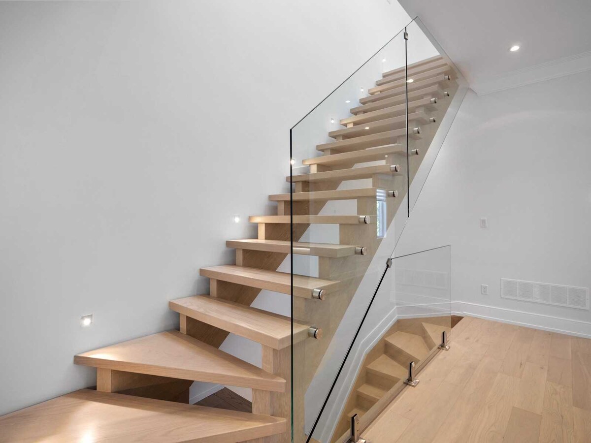 A modern staircase features light-colored wooden steps complemented by sleek glass railings. Soft lighting accentuates the elegant design, while the walls remain painted in a neutral tone, enhancing the contemporary feel of the space.