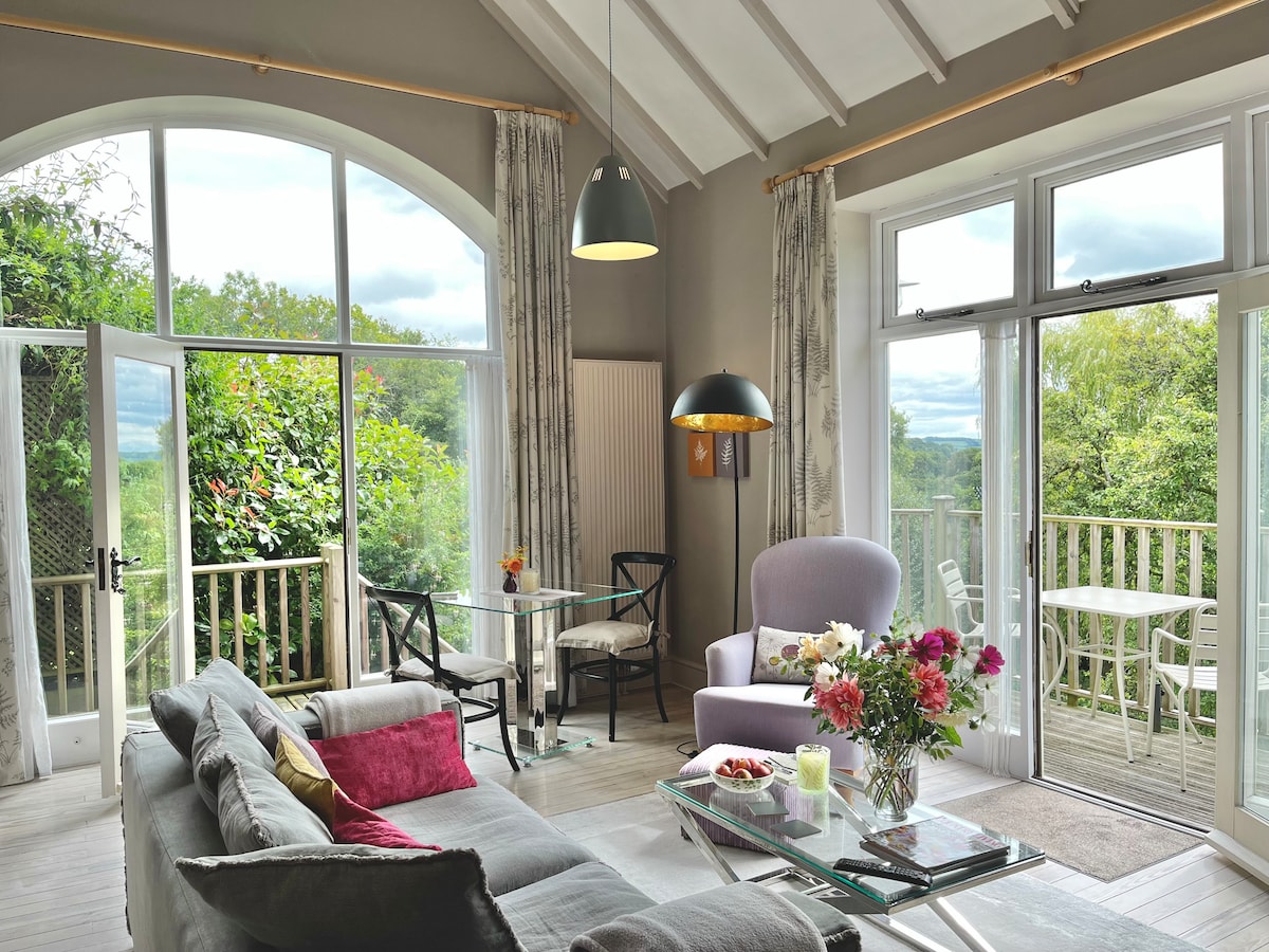 A bright sitting area features large windows that invite natural light and showcase views of the surrounding greenery. Comfortable seating includes a light purple armchair and a gray sofa adorned with cushions. A glass dining table is set for two, complemented by fresh flowers.