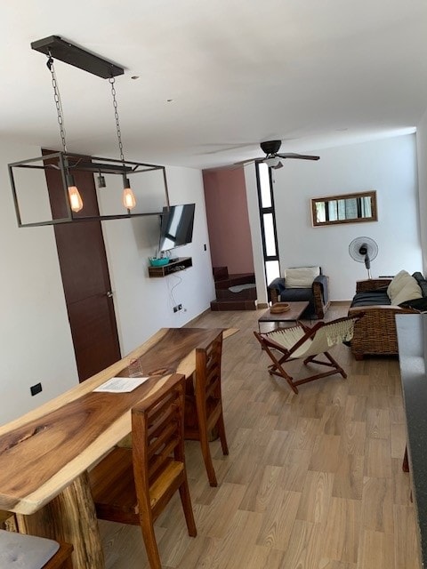 An open living area features a wooden bar counter with seating for four. A wall-mounted TV is positioned opposite a comfortable seating arrangement that includes a fan and a lounge chair. Natural light is invited through large windows, enhancing the spacious feel of the area.
