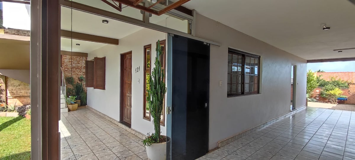 A corridor is visible, showcasing a tiled floor and neutral-colored walls. Doors lead to individual rooms, with potted plants positioned along the path. Windows allow natural light to fill the space, enhancing the open and airy feel of the area.