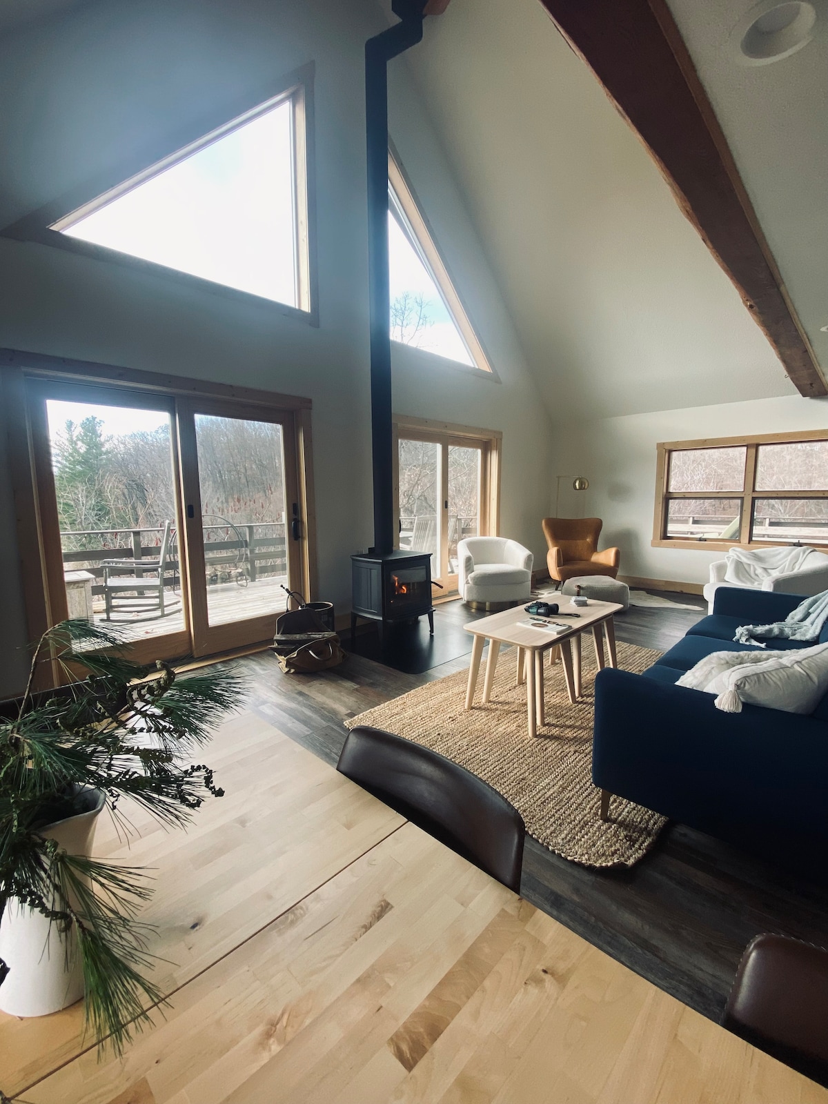 A spacious living area features vaulted ceilings and large triangular windows that invite natural light. A modern wood stove provides warmth, while cozy seating is arranged around a wooden coffee table. A woven rug adds texture, complementing the wooden flooring that extends throughout the space.