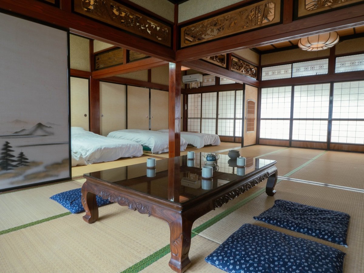 A spacious traditional Japanese room features tatami flooring and sliding doors with artwork. Two beds are arranged on one side, and a wooden table sits in the center, surrounded by blue floor cushions. Natural light filters through the shoji windows, enhancing the serene ambiance.