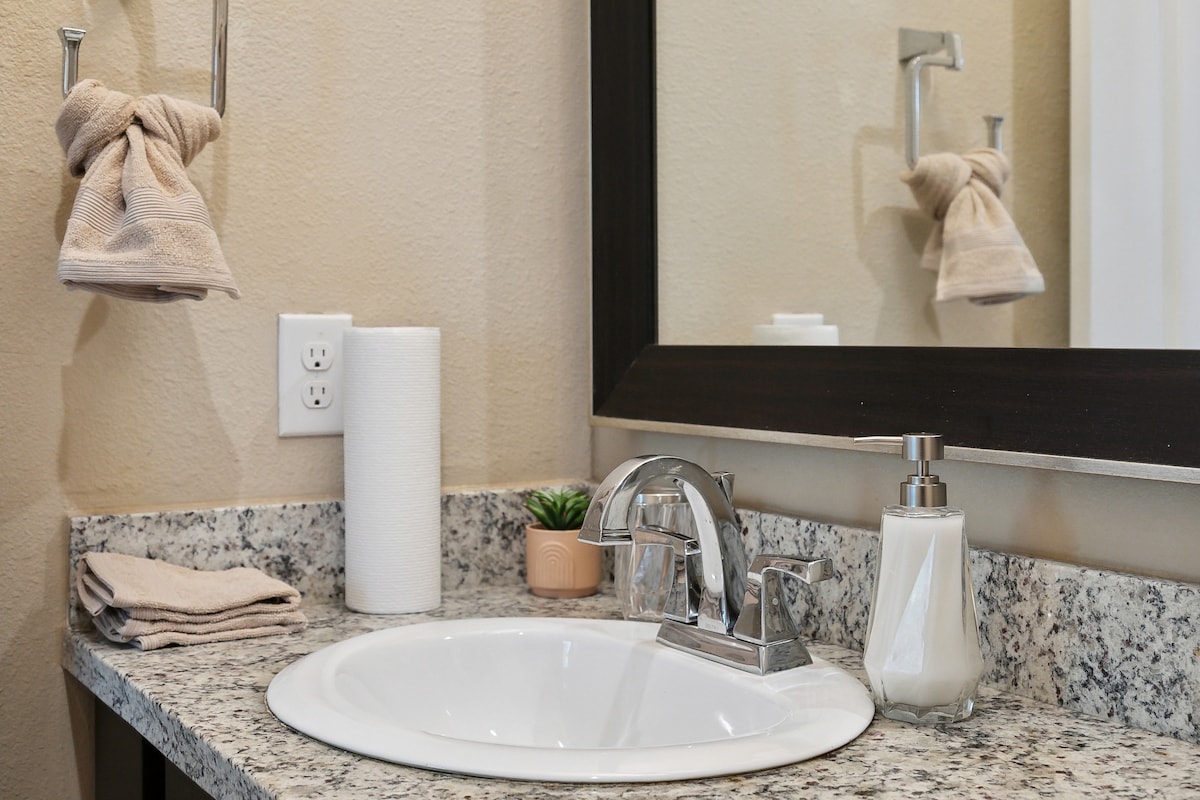 A clean bathroom countertop is shown, featuring a round sink with a modern faucet. Two beige hand towels are hung neatly on the side. A small plant and a dispenser for liquid soap are placed next to a roll of paper towels. A large mirror reflects the space.