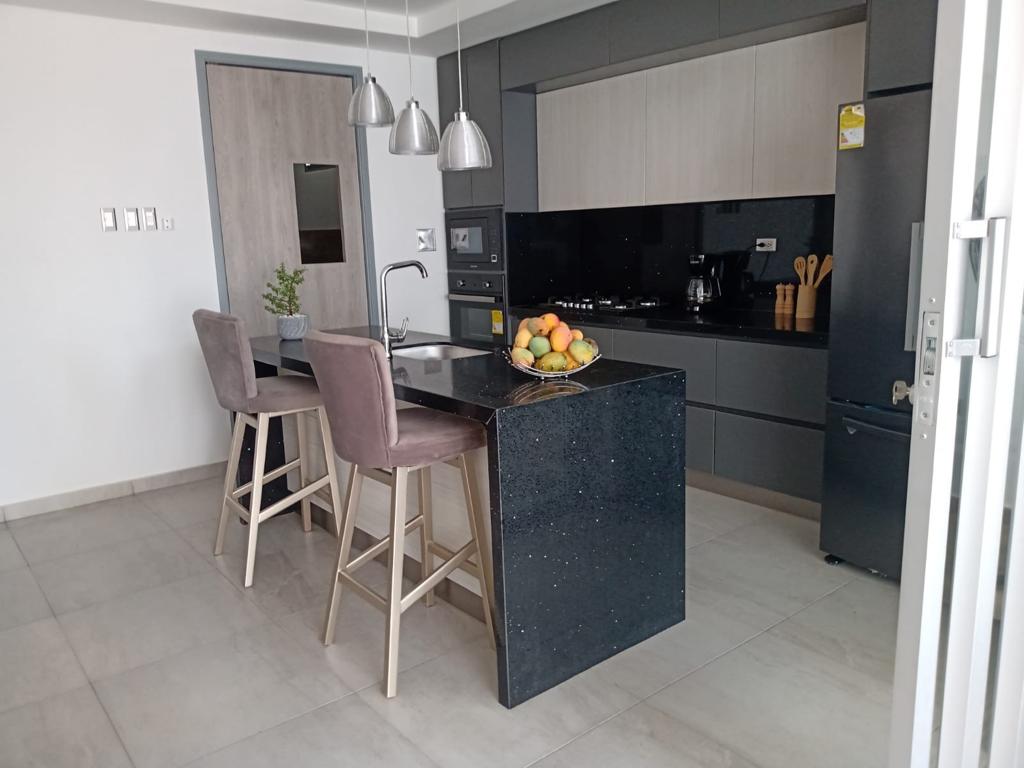 A modern kitchen features sleek cabinetry and a dark countertop with a fruit bowl. Two upholstered barstools are positioned at a spacious island, while pendant lights provide illumination above. Appliances are integrated seamlessly into the design, creating an inviting cooking space.