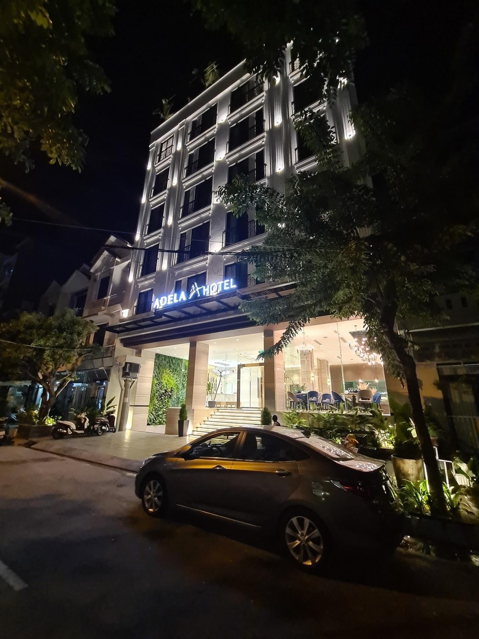 The hotel exterior is elegantly illuminated at night, showcasing modern architectural features. A sleek car is parked nearby, while lush greenery surrounds the entrance, providing a welcoming atmosphere. The entrance is adorned with well-lit signage, enhancing visibility and creating a sense of openness.