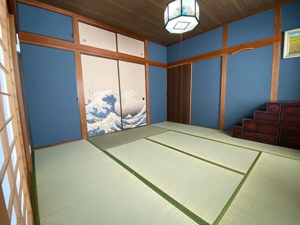 A traditional tatami room is presented, featuring blue walls and sliding doors adorned with a wave motif. A modest light fixture hangs from the ceiling. The room's spaciousness is enhanced by natural light, casting a serene ambiance across the tatami flooring.
