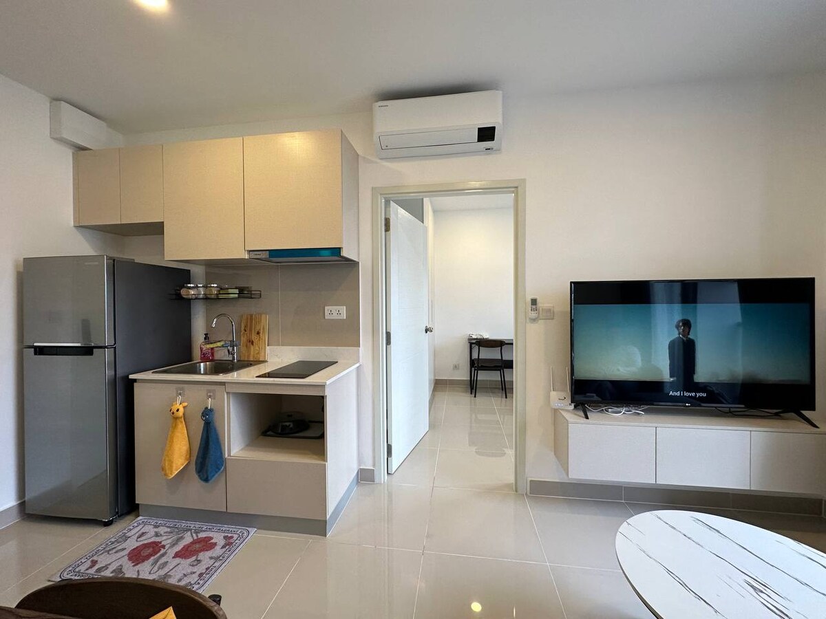 A well-equipped kitchen area is visible, featuring modern appliances including a fridge and a stove. A seamless passthrough connects to a living space with a television. Light tiles create a bright atmosphere, and a small decorative rug adds a touch of comfort.