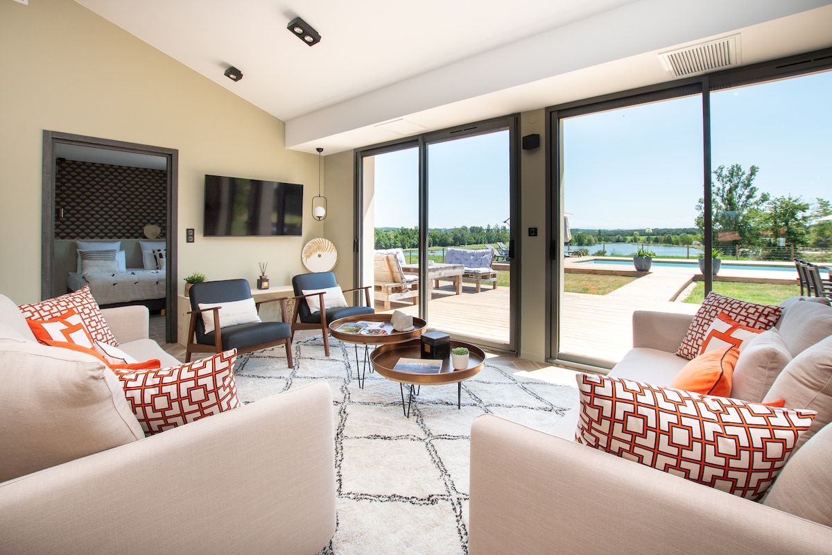 A modern living area features large glass doors that open to a scenic outdoor space, with views of the pool and surrounding greenery. Comfortable sofas are arranged around a coffee table, complemented by stylish decor and natural light pouring in.
