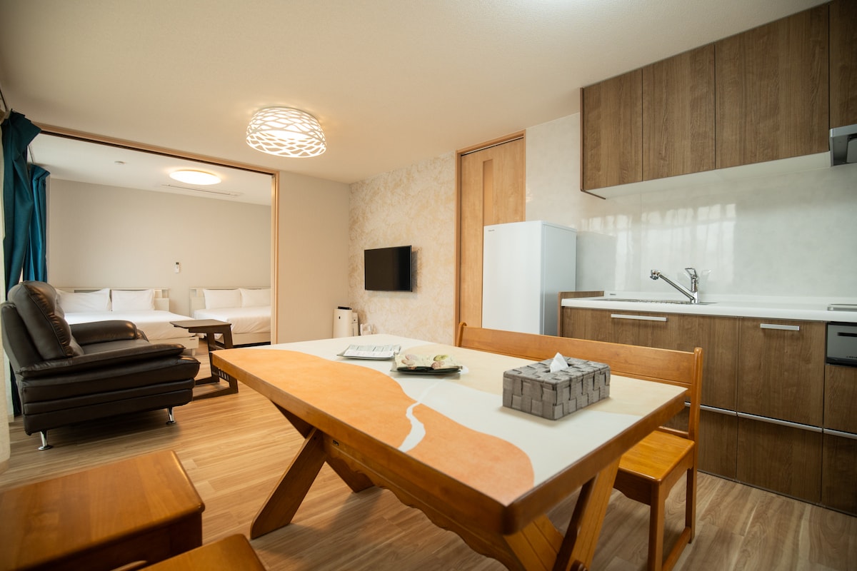 A dining area is presented with a wooden table and chairs, inviting communal meals. An open kitchen area is visible, featuring cabinetry and essential appliances. The living space in the background offers a cozy couch and a mounted television, creating a comfortable area for relaxation.
