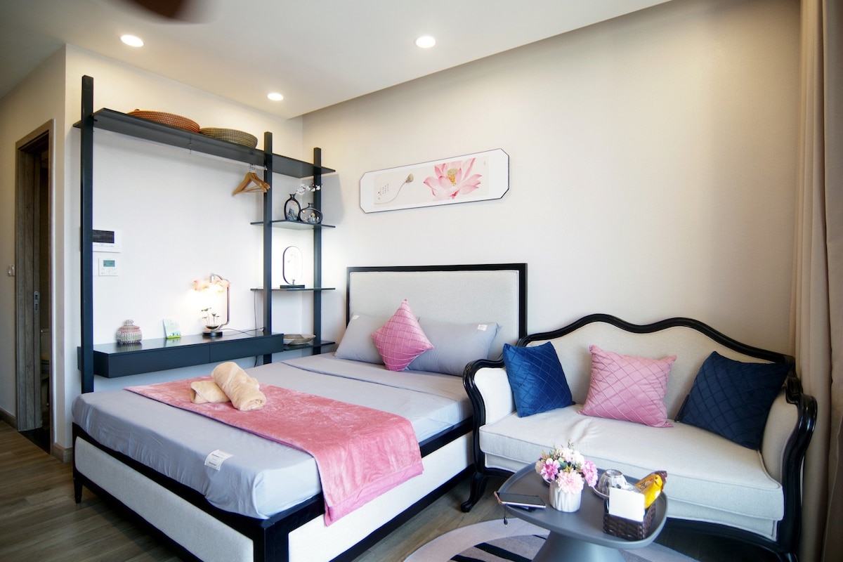 A comfortable sleeping area features two spacious beds dressed in soft linens. A plush sofa with patterned cushions complements a small coffee table arranged with decorative items. Natural light fills the room through a nearby window, enhancing the modern decor.