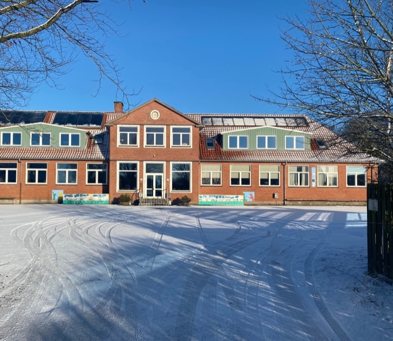 The exterior of a charming brick building is shown, featuring large windows and a symmetrical facade. A snow-covered ground surrounds the structure, and clear blue skies provide a bright backdrop. The building reflects its history as a former village school, with details hinting at its unique character.