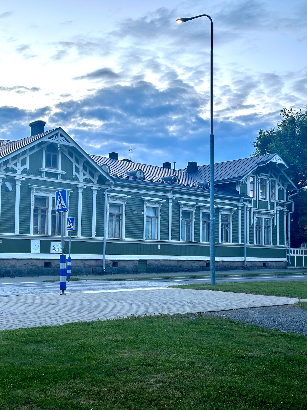 The image depicts a historical wooden building with a green facade and white trim. The building features intricate architectural details, including dormer windows and a steep roof. A paved pathway and grassy area are visible in the foreground, along with a lamppost and street signs.