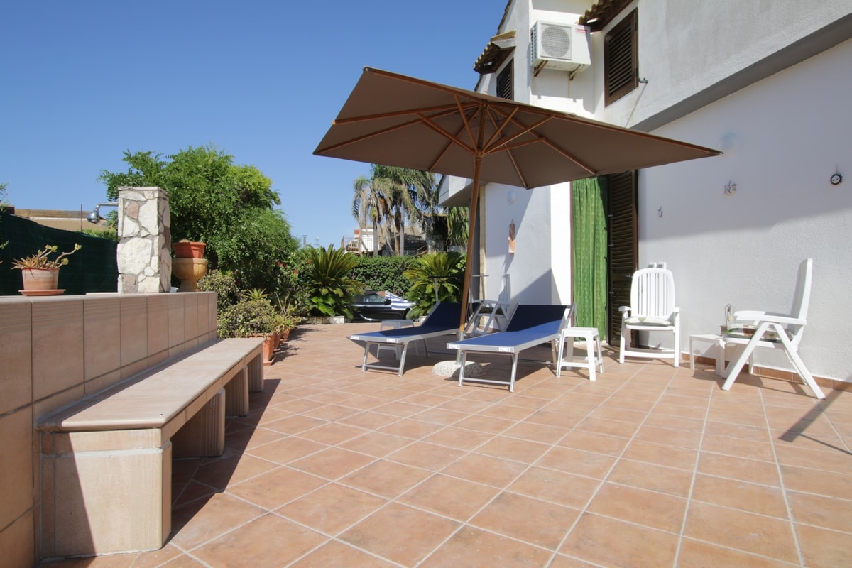 An outdoor terrace is presented with two lounge chairs positioned under a large umbrella. A stone bench runs along one side, surrounded by potted plants and greenery. The space is designed to provide a relaxing atmosphere, with ample sunlight filling the area.