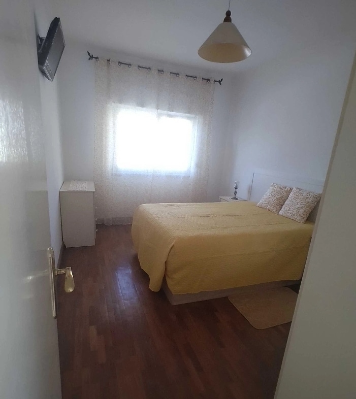 A bright sleeping area features a queen-sized bed dressed in a pale yellow coverlet. Natural light enters through a sheer-curtained window, illuminating the warm wood flooring. A small bedside lamp and a white bedside table are positioned next to the bed.