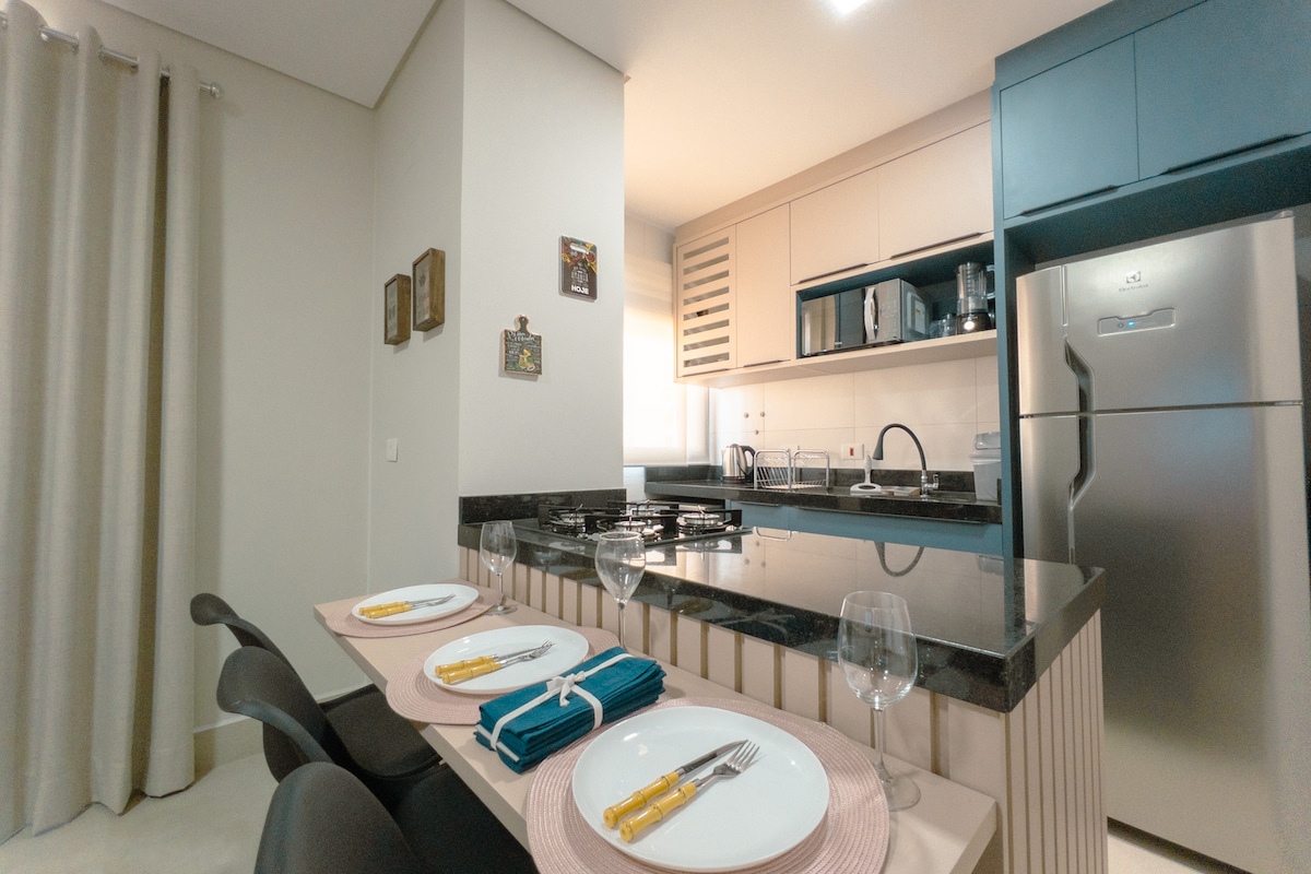 A modern kitchen area features a sleek countertop with dining settings for four. A well-equipped kitchen is visible, complete with an oven, a microwave, and a refrigerator. Natural light enters through a nearby window, while neutral curtains frame the space.